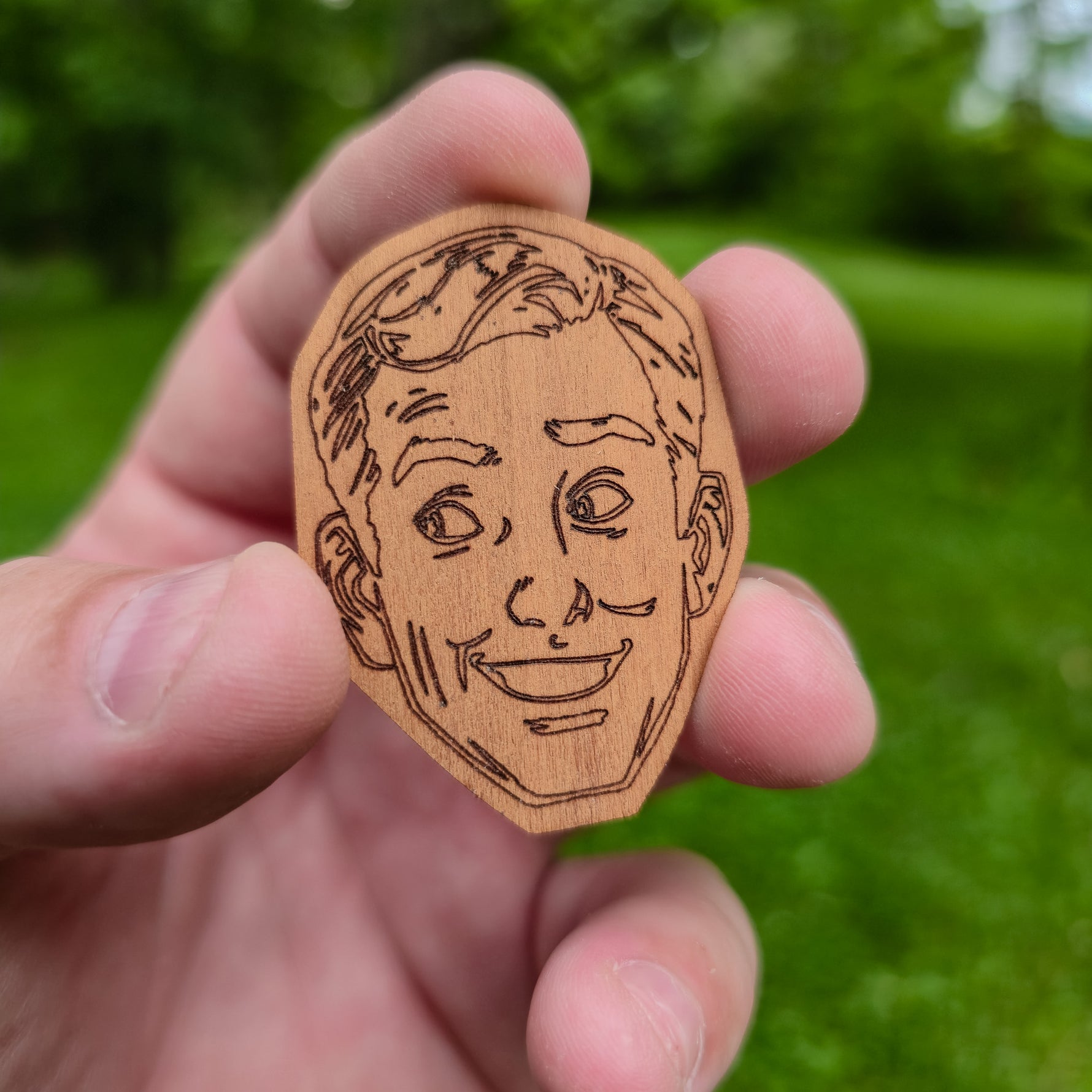 Wooden sticker with a engraved face held in a hand against a green outdoor background