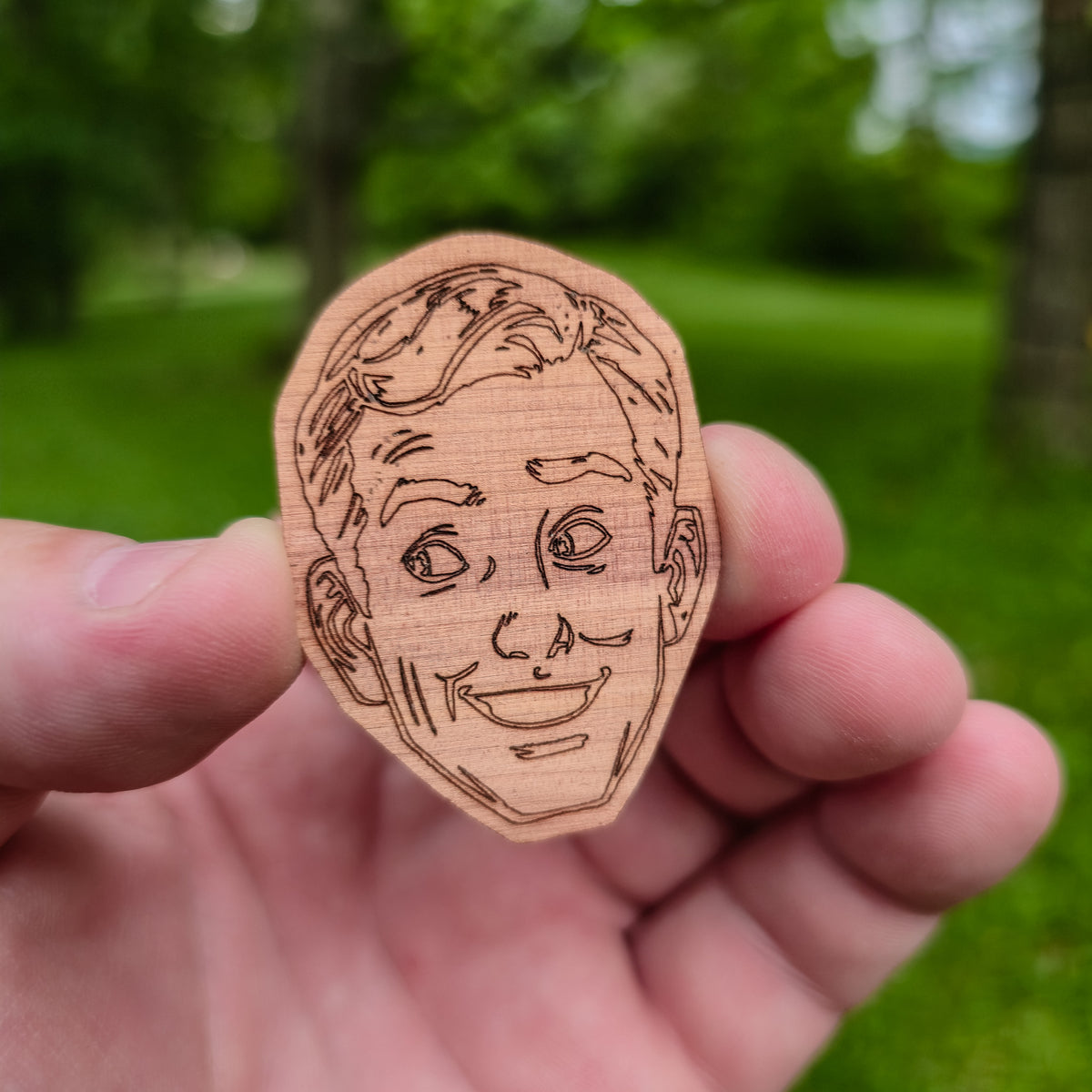 Wooden sticker of a face held in a hand with a green outdoor background