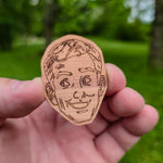 Wooden sticker of a face held in a hand with a green outdoor background