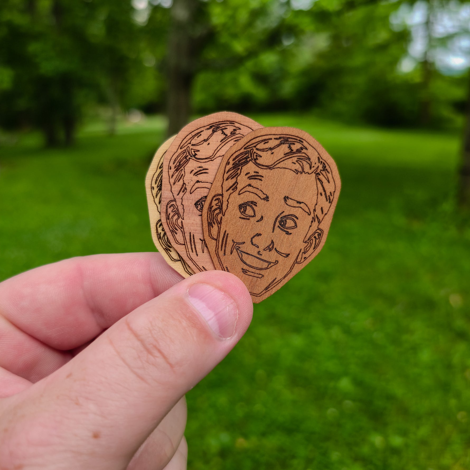Three wooden stickers with engraved faces held against a green outdoor background