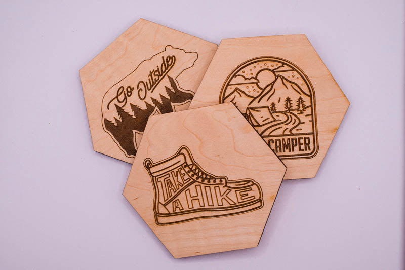 Three wooden coasters with engraved designs on a light purple background