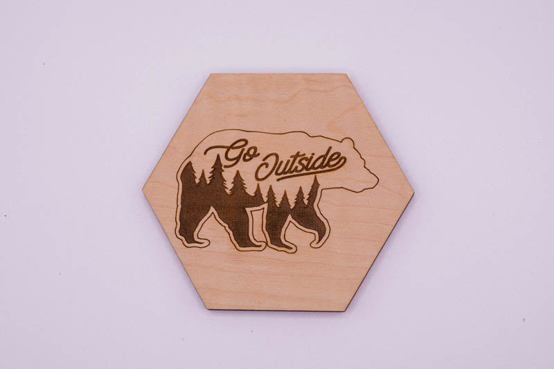 Hexagonal wooden coaster with bear design and 'Go Outside' text on a light purple background