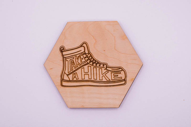 Hexagonal wooden coaster with a laser-cut shoe design and 'Take a Hike' text on a light purple background.
