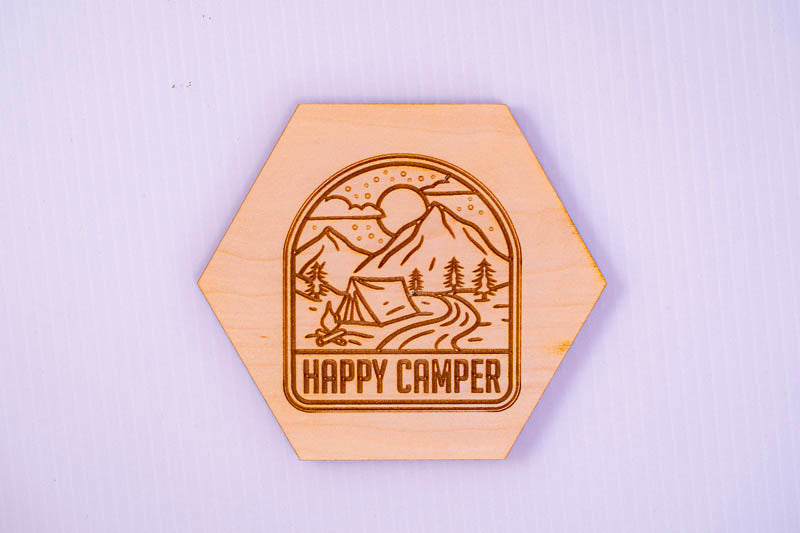 Hexagonal wooden coaster with 'Happy Camper' design on a light purple background