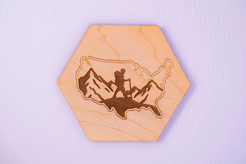 Hexagonal wooden coaster with engraved design of a person hiking in the mountains on a light purple background