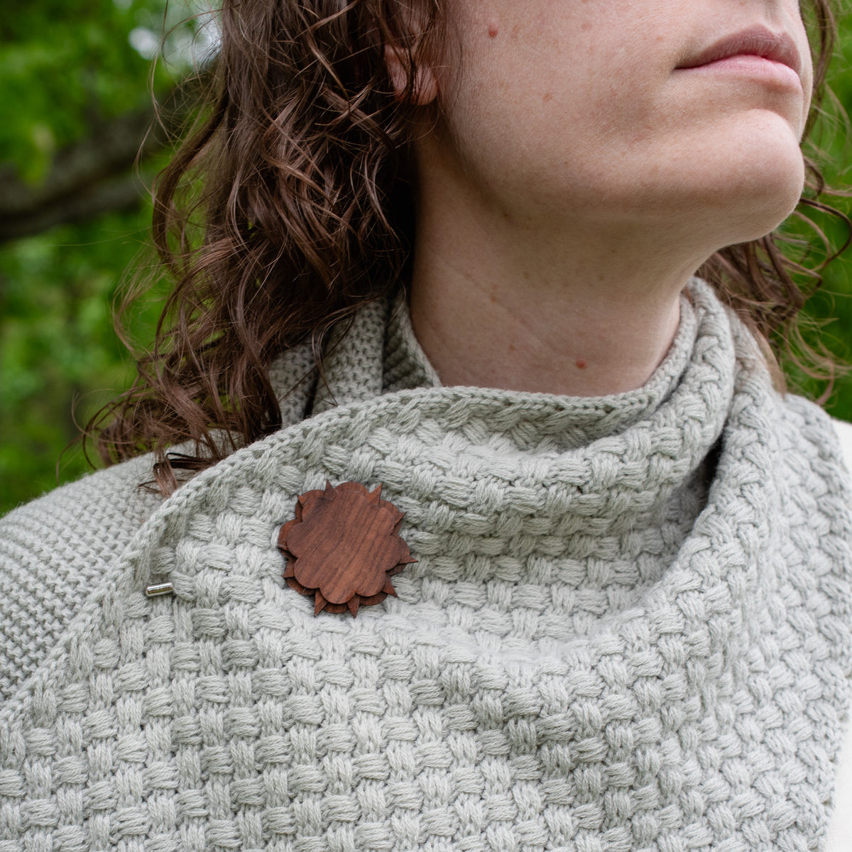Person wearing a textured garment with a wooden brooch, surrounded by greenery.