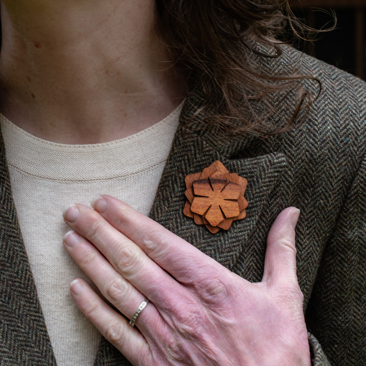 Person wearing a brown textured jacket with a wooden brooch, holding their hand to chest level.