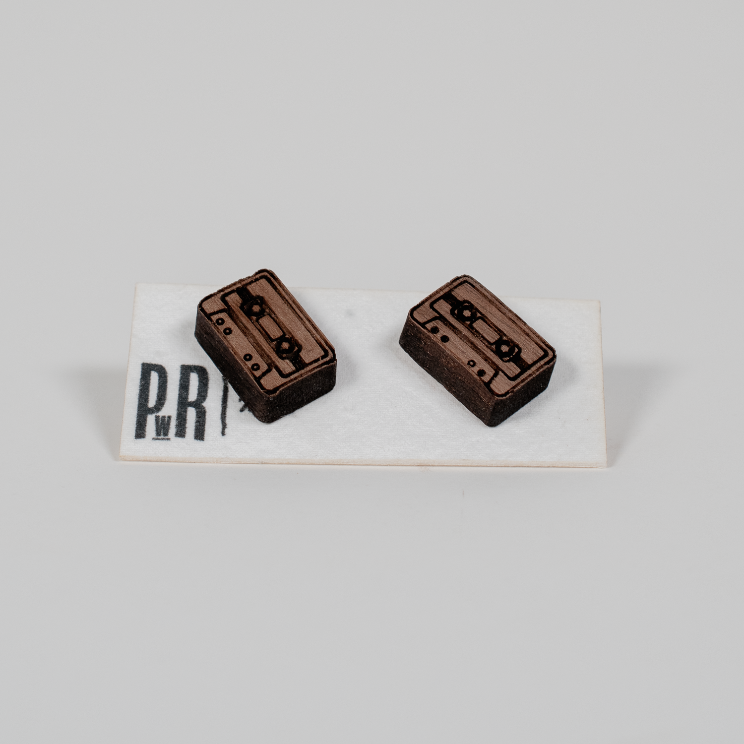 Pair of small laser-engraved wood stud earrings with geometric cassette tape design, natural finish, displayed on branded card.