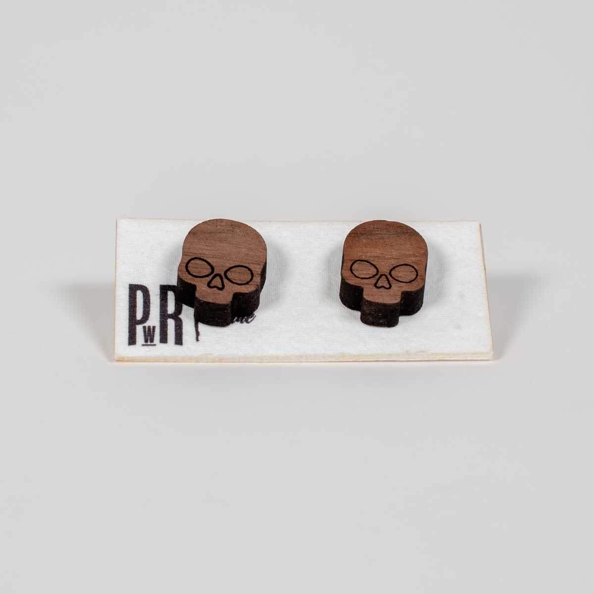 Small wooden skull stud earrings with geometric engraved design, natural wood finish, displayed on a branded PRW Made white card.