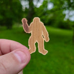Hand holding a brown Bigfoot silhouette sticker against a green outdoor background