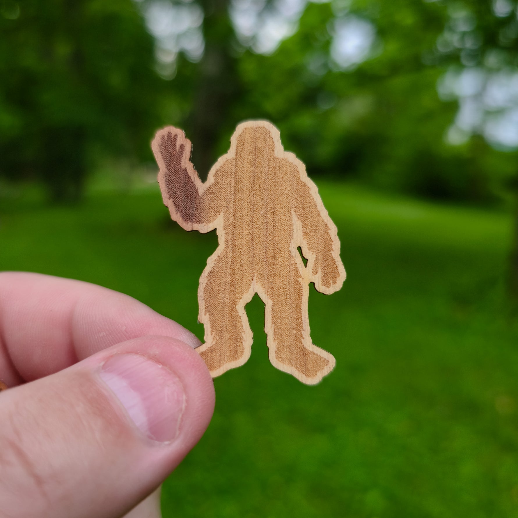 Hand holding a brown Bigfoot silhouette sticker against a green outdoor background