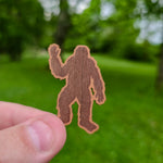Hand holding a brown sticker of a bigfoot silhouette against a blurred green outdoor background