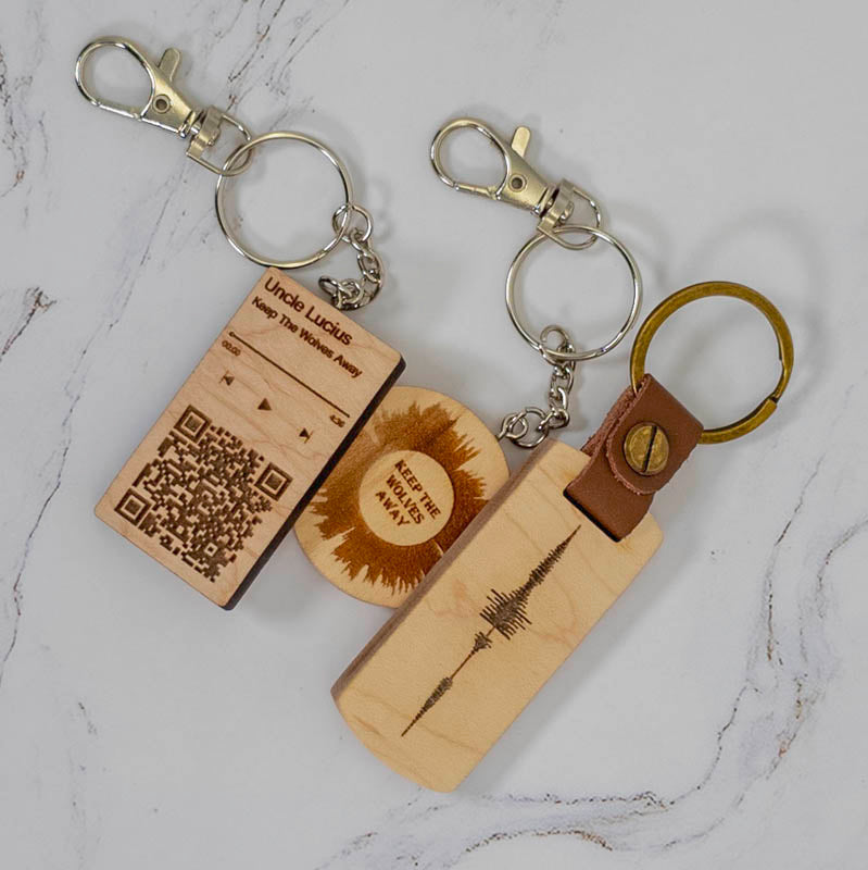 Three custom wooden keychains in maple - a Spotify music QR code, a sunburst with custom text, and a sound wave on a leather and brass fob - laser engraved by PRWMade