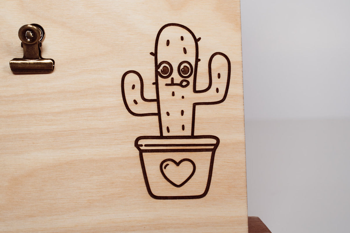 Detail of a line art cactus in a heart-shaped pot laser engraved on a light natural wood message board by PRWMade