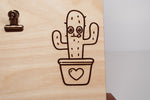 Detail of a line art cactus in a heart-shaped pot laser engraved on a light natural wood message board by PRWMade