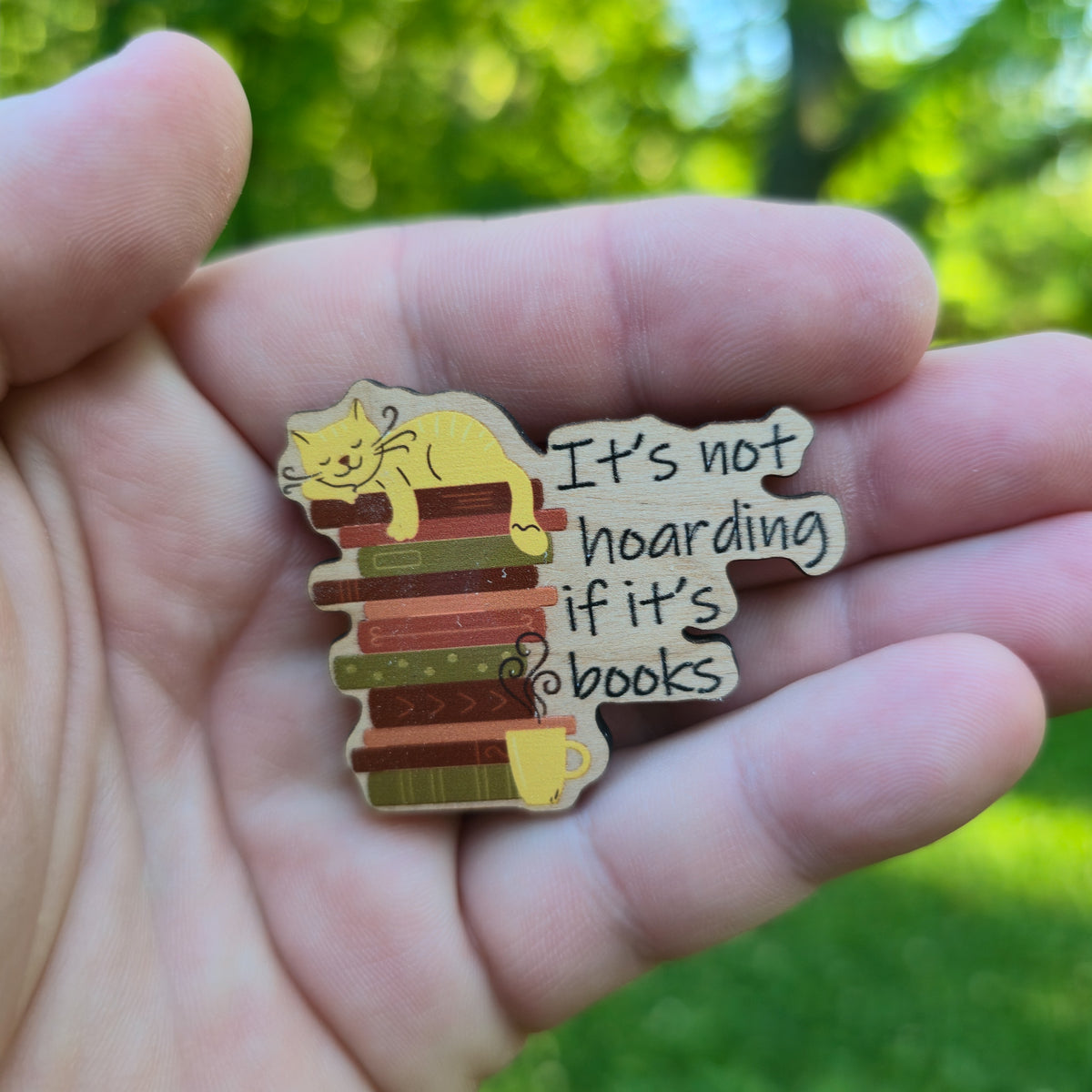 Wooden magnet with a cat on books design and text 'It's not hoarding if it's books' held in a hand with a green blurred background.