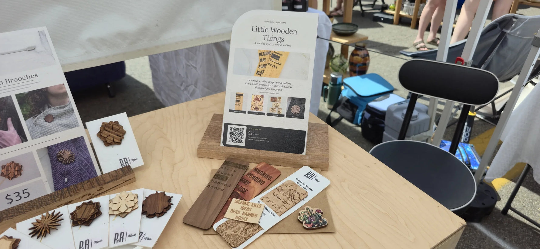 Little Wooden Things subscription display at a craft fair booth showing the promo card, bookmarks, brooches, and stickers included in the monthly mystery mail