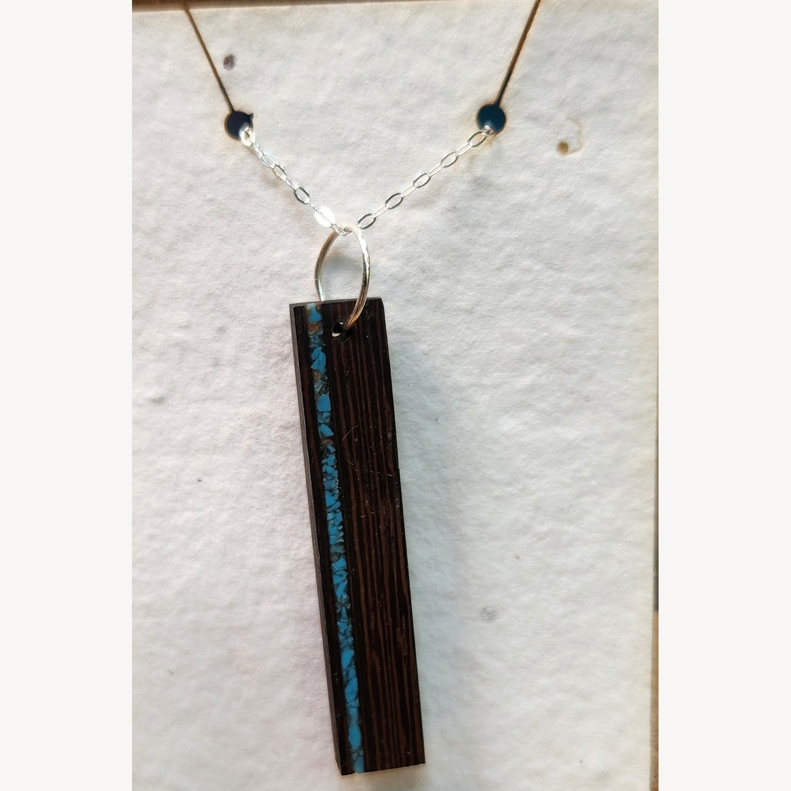 Wenge bar pendant with a vein of crushed turquoise inlay along one long edge, hanging from a sterling silver chain on a white seed-paper display card