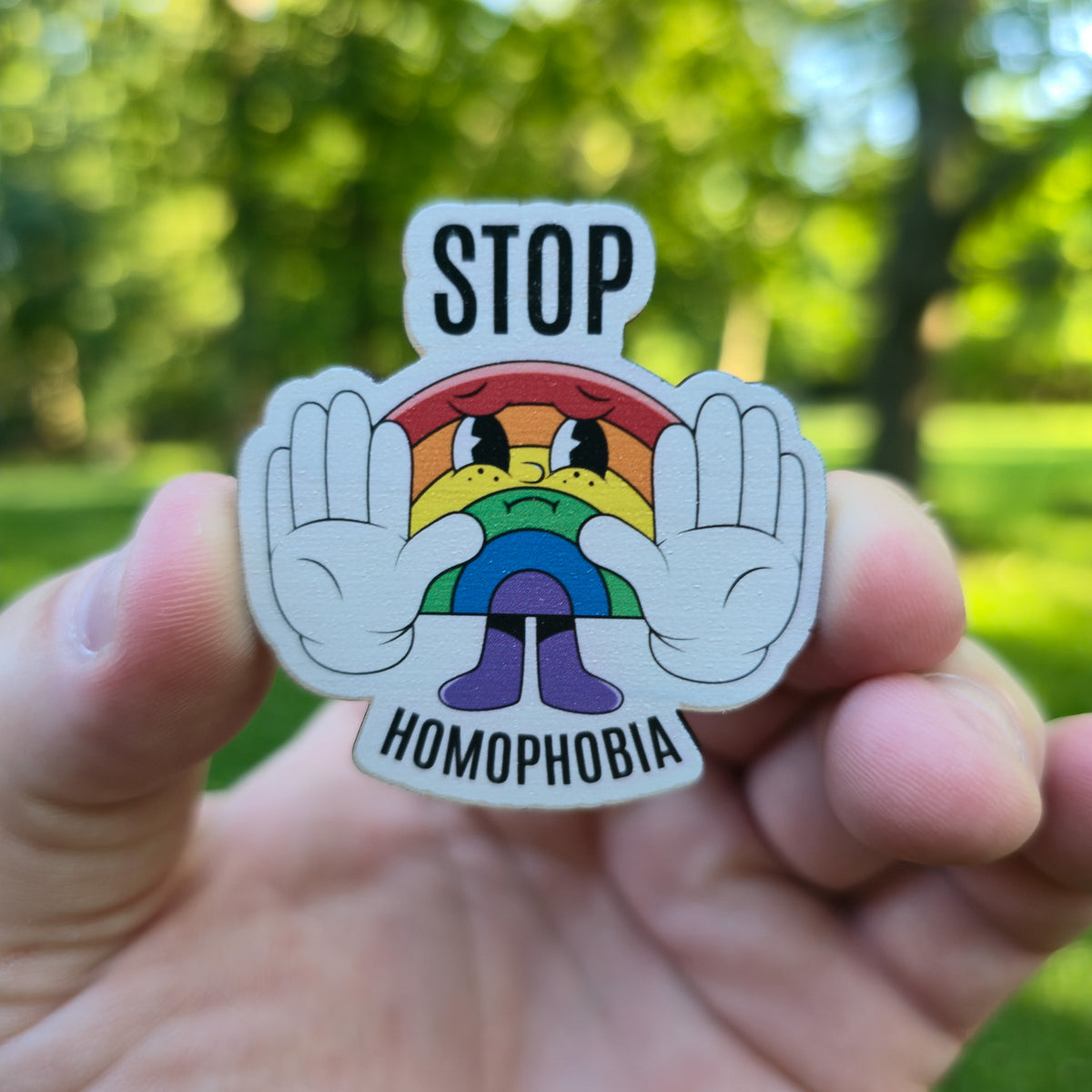 Magnet with rainbow character and 'Stop Homophobia' text held in a hand against a blurred green outdoor background