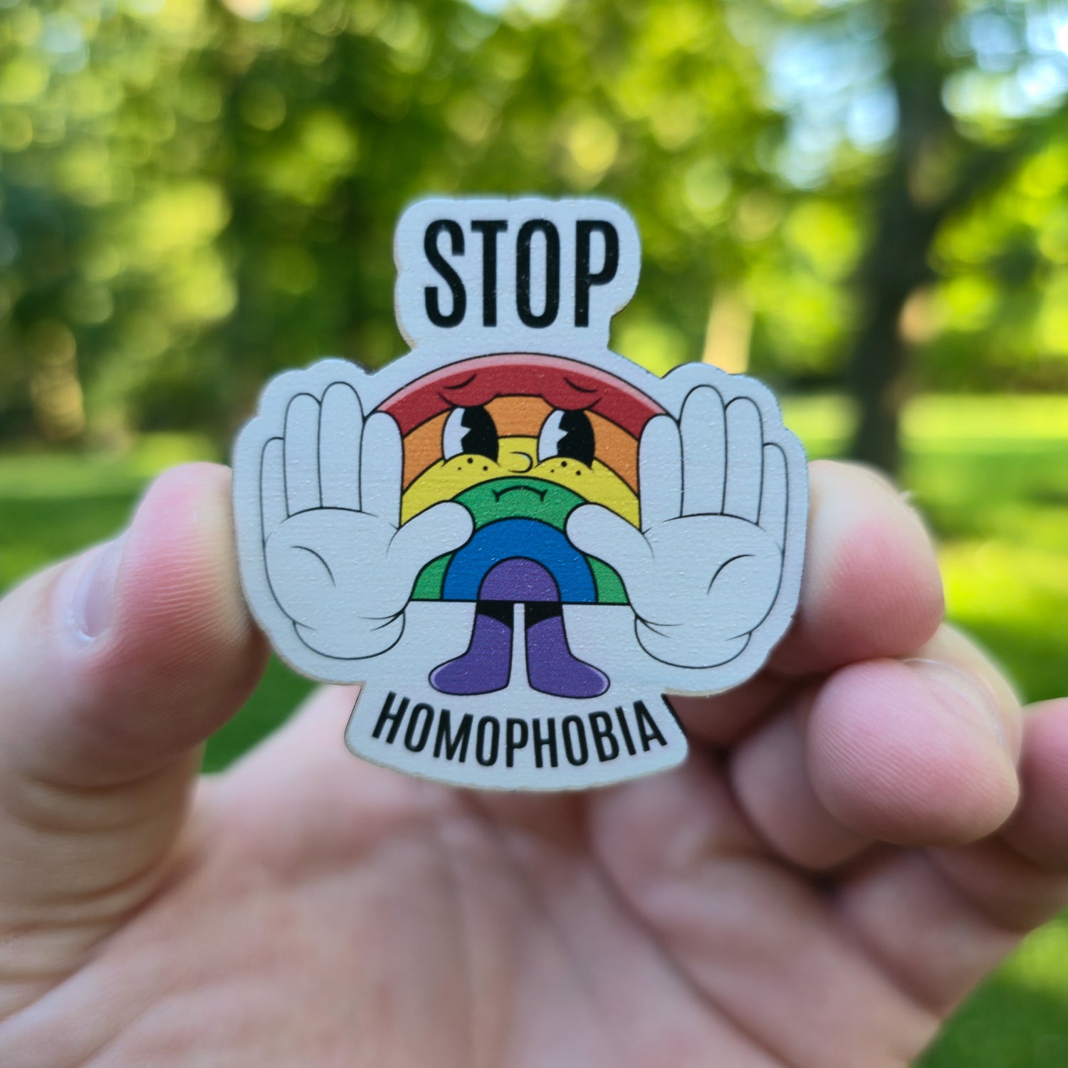 Magnet with rainbow character and 'Stop Homophobia' text held in a hand against a blurred green outdoor background