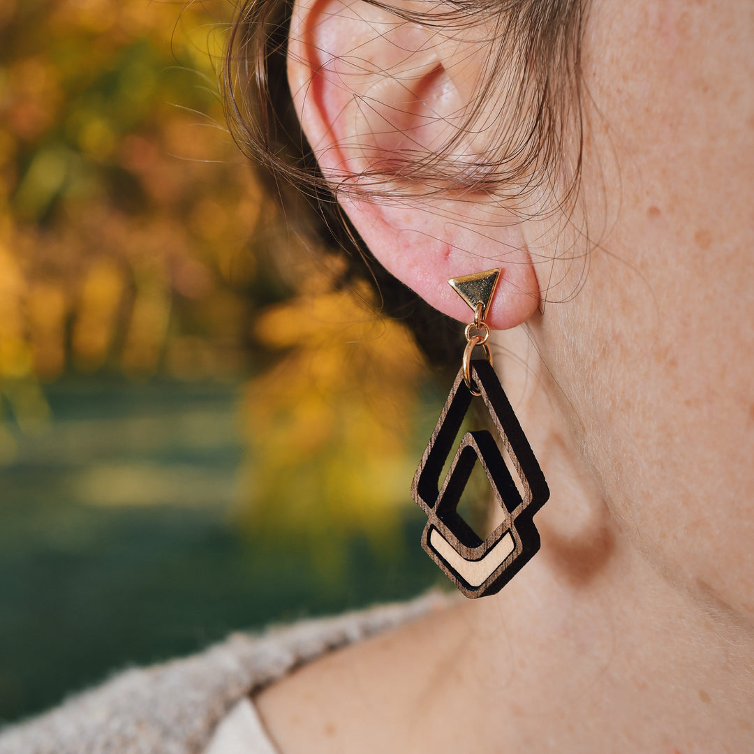 A close-up of a person's ear wearing a geometric wooden dangle earring. The earring features layered triangular and chevron shapes in natural wood with dark brown painted edges, set against a blurred outdoor background of green and yellow foliage.