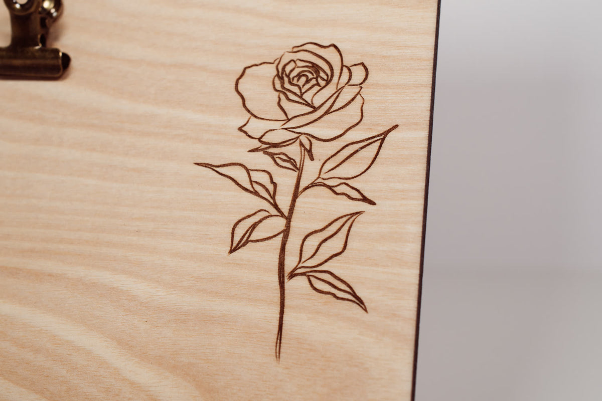 Detail of a laser engraved rose with stem and leaves on a natural wood message board, fine line engraving by PRWMade